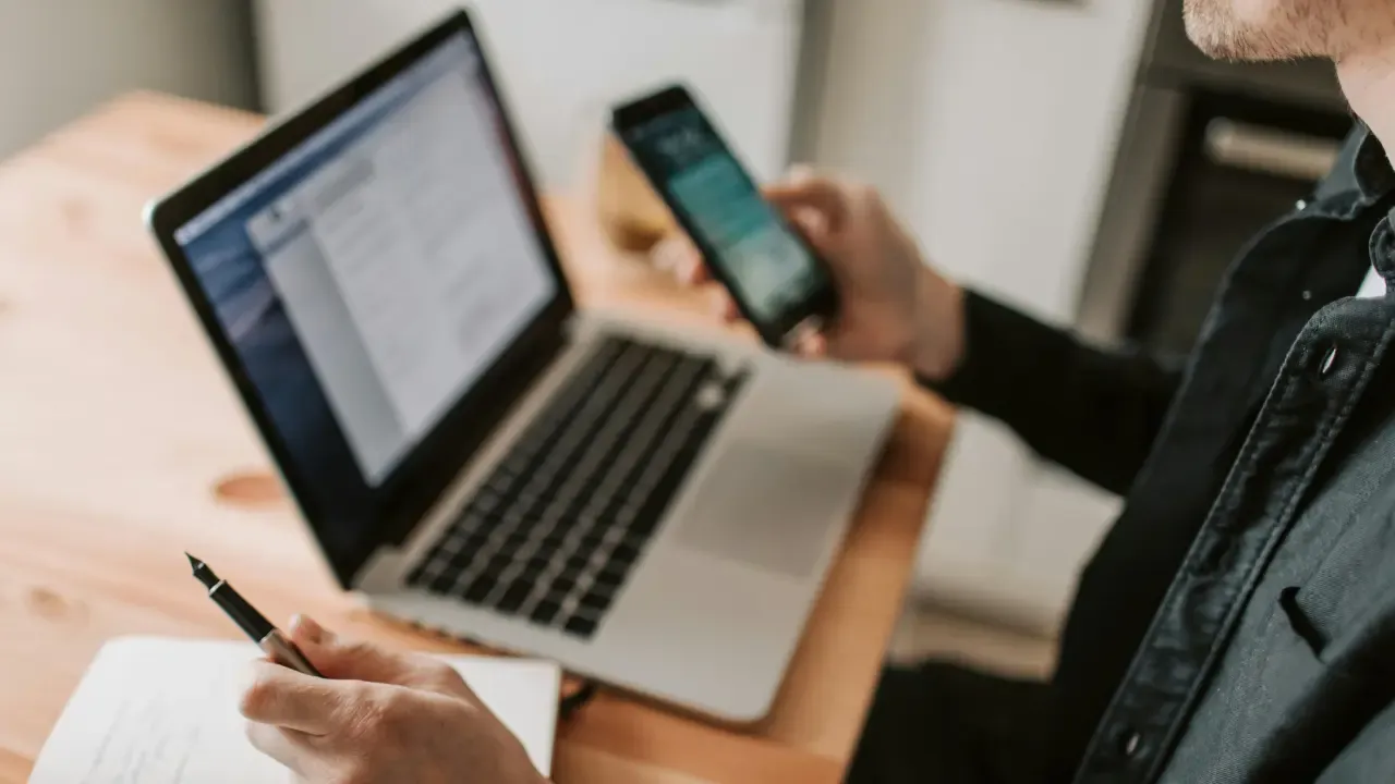 En la foto: Hombre con celular en la mano, de fondo un computador portátil. 